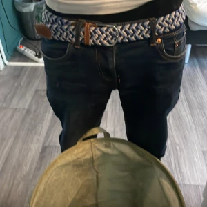 Blåvit flätad bälte med läderdetaljer - Säljer ett snyggt flätat bälte i blått och vitt med bruna läderdetaljer vid spännet. Bältet har ett silverfärgat metallspänne och passar perfekt till jeans. Stilen är sportig och modern, och bältet är flexibelt tack vare den flätade designen.