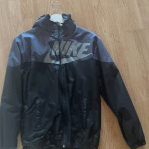Svart Nike vindjacka Dri-Fit M - Nike vindjacka i svart med stor logga över bröstet. Jackan har huva, dragkedja framtill och två fickor. Tillverkad i lätt och sportig polyester med Dri-Fit-teknologi. Perfekt för dig som gillar en clean och sportig stil. Köpt i Vietnam, liten i storlek. Mer som S skulle jag säga 