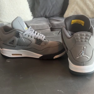 Nike Air Jordan 4 Cool Grey sneakers - Nike Air Jordan 4 Cool Grey med grå mocka, vita och svarta detaljer samt gula accenter på plösen. Klassisk meshpanel på sidorna, Jumpman-logga bak och på plösen. Skorna har snörning och en bekväm, vadderad insida. Perfekta för dig som gillar streetwear och sneakerskultur.