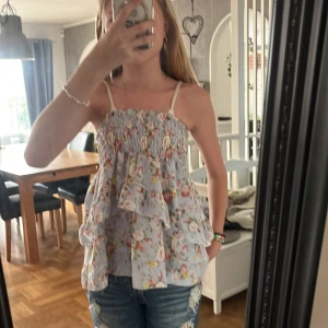 Blommig volangtop med smock S/M - Supersöt blommig topp med volanger och smock upptill. Ljusblå botten med rosa, gula och gröna blommor. luftig passform, perfekt för varma dagar. Materialet är en mix av bomull och polyester. Kan även användas som en kjol.