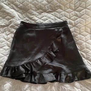 Svart sidenkjol med volang Gina Tricot - Supersnygg svart kort omlottkjol från Gina Tricot med volangdetaljer längs nederkanten. Kjolen har en glansig satinliknande yta och resår i midjan för skön passform. Perfekt för dig som vill ha en trendig och stilren look.