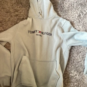 Ljusgrå hoodie från Tommy Hilfiger - Snygg ljusgrå hoodie från Tommy Hilfiger med klassisk logga broderad på bröstet. Tröjan har huva med snörning och en stor magficka framtill. Perfekt för en chill och avslappnad stil.