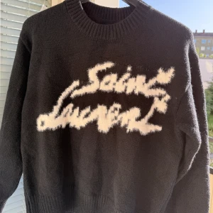 Svart Saint Laurent stickad tröja - Svart stickad tröja från Saint Laurent med vit, fluffig logotyp framtill i en cool, handskriven stil. Tröjan har rund halsringning, långa ärmar och ribbade muddar. Perfekt för dig som vill ha en statement piece med lyxig känsla.