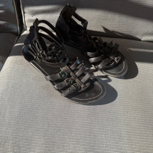Svarta gladiatorsandaler med dragkedja - Svarta gladiatorsandaler i skinnimitation med flera remmar och snygga metallspännen. Dragkedja baktill för enkel påtagning och blommönster under sulan. Perfekta för en cool sommarlook.