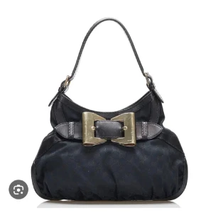 Svart handväska från Gucci med rosett - Snygg svart handväska från Gucci med en stor dekorativ rosett i guldmetall framtill. Väskan har en klassisk bågformad siluett, kort axelrem och är tillverkad i skinn och textil. Perfekt för dig som gillar statement-detaljer och lyxig känsla. Köpt secondhand men jätte fint skick. Skriv om ni har frågor💛