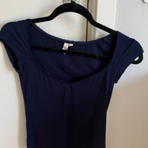 Mörkblå topp med v-ringning - En enkel och stilren mörkblå topp med v-ringad hals och korta ärmar. Toppens material känns mjukt och stretchigt, perfekt för en avslappnad look. Liten spetsdetalj längs halsringningen ger en söt touch.