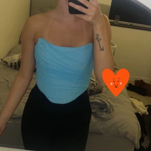 Ljusblå korsett tubtopp - Säljer en snygg ljusblå korsett tubtopp med draperad effekt. Toppen är axelbandslös och har en tight passform som framhäver figuren. Perfekt för dig som vill ha en stilren och trendig look.