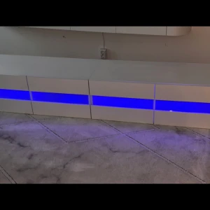 Tv bänk  - Bilderna visar en modern TV-bänk eller förvaringsmöbel med vit yta och en blå LED-ljusrand. 180cm lång. Skriv vid funderingar eller frågor! 