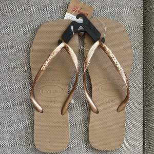Beige och roséguld Havaianas sandaler - Snygga Havaianas sandaler i beige med metallic roséguldfärgade remmar och fyrkantig tå. Klassisk flip-flop-modell med logga på remmen, perfekt för sommardagar. Tillverkade i syntetmaterial för en lätt och bekväm känsla. Har en svart fläck, men det är inget som syns vid användning❤️