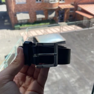 Svart klassiskt skärp i skinn - Säljer ett stilrent svart bälte i äkta skinn med en enkel, fyrkantig silverspänne. Bältet är slätt utan mönster och passar perfekt till jeans eller kostymbyxor. Tidlös design som funkar till många olika outfits.