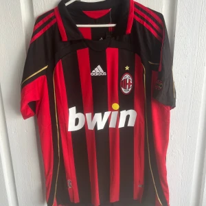 AC Milan Kaka' 22 Adidas matchtröja - Säljer en klassisk AC Milan fotbollströja från Adidas med Kaka' och nummer 22 på ryggen. Tröjan är röd och svart med guldiga detaljer, Champions League-märke på ärmen och Bwin som sponsor på bröstet. Ikoniska ränder och klubbmärke framtill.