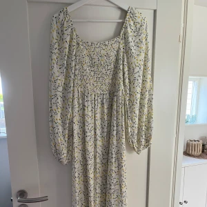 Lång blommig klänning med smock - Superfin långklänning med blommigt mönster i vitt, gult och svart. Klänningen har smockad överdel, långa puffärmar och en luftig passform. Perfekt för sommarens alla tillfällen och ger en romantisk vibe.