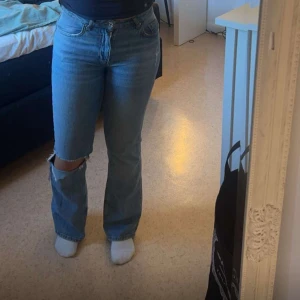 Ljusblå bootcut jeans med hål - Säljer ett par ljusblå jeans med bootcut-modell och ett stort, slitet hål över ena knät. Jeansen har klassisk femficksdesign, hög midja och är i ett mjukt denimtyg. Perfekta för en avslappnad och trendig look.