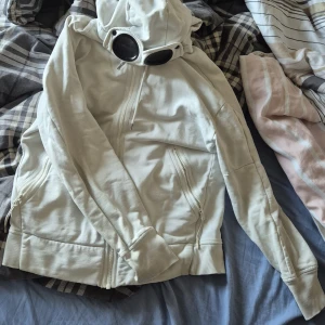 Vit Goggle-hoodie från C.P. Company - Säljer en clean vit hoodiejacka från C.P. Company med ikoniska goggles i huvan. Jackan har dragkedja framtill, två snedställda fickor med dragkedja och ribbade muddar. Perfekt för dig som vill sticka ut med streetwear-vibe och unik design. Det är size m men passar både i s och xs ör 175cm och passar perfekt. Min kompis är 165cm och den passar honom också bra. Äkta pris 2899kr. Mitt pris 1399kr