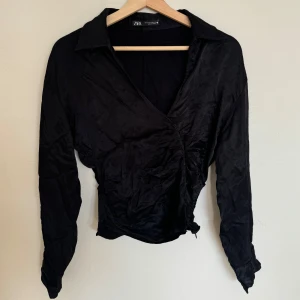 Svart omlottopp från Zara - Svart omlottopp från Zara med krage och långa ärmar. Toppen har en lätt glansig yta och knytning i sidan.