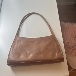 Brun handväska i skinn från Gucci - Säljer en snygg brun handväska från Gucci i äkta skinn. Väskan har en stilren, avlång form med två smala axelband och diskret Gucci-logga präglad framtill. Invändigt finns flera fack och dragkedja för smart organisering. Perfekt för dig som gillar klassisk och tidlös design.