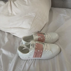 Valentino Rockstud sneakers vit/rosa - Snygga Valentino Rockstud sneakers i vitt skinn med ljusrosa detaljer och ikoniska nitar. Är använd max 2-3 gånger köpte begagnande så vet ej till hundra procent att dem är riktiga fick ett kvitto med köptet som tyder på att dem är riktiga men är inte 💯 Skorna har platt sula, rund tå och vita snören. Perfekta för dig som vill sticka ut med en lyxig och trendig touch.