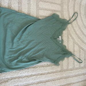 Grön ribbad linnetopp med spets - Snygg grön linnetopp med tunna axelband och v-ringning dekorerad med spets. Toppen är ribbad och har en mjuk, stretchig känsla som gör den superbekväm. Perfekt att styla med jeans eller kjol för en chill look.