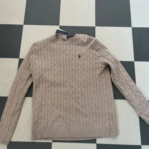 Beige kabelstickad tröja Polo Ralph Lauren - Kabelstickad tröja i beige/aprikos från Polo Ralph Lauren. Klassisk rund halsringning, ribbade muddar och diskret broderad logga på bröstet. Perfekt för lager-på-lager och snygg till både jeans och kjol.