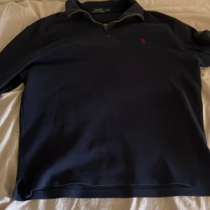 Mörkblå half zip tröja Polo Ralph Lauren - Mörkblå långärmad tröja från Polo Ralph Lauren med klassisk half zip-dragkedja och röd broderad logga på bröstet. Tröjan har en stilren krage och är tillverkad i mjuk bomull, perfekt för lager-på-lager under kyliga dagar. 