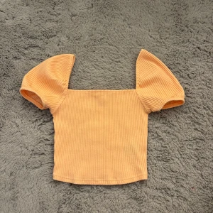 Ribbad persikofärgad topp med puffärm - Superfin ribbad topp i persikofärg med fyrkantig halsringning och korta puffärmar. Croppad modell som ger en trendig vibe, perfekt till höga jeans eller kjol. Mjuk och stretchig för skön passform.