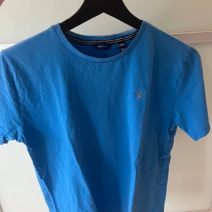 Blå t-shirt från GANT - Snygg blå t-shirt från GANT med klassisk rund hals och liten broderad logga på bröstet. T-shirten är i mjuk bomull och har en enkel, stilren look som passar perfekt till jeans eller shorts.