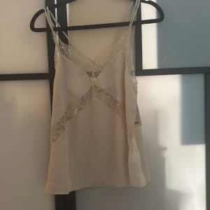 Champagnefärgad linnetopp från BikBok - Supersöt champagnefärgad linnetopp från BikBok med tunna axelband och v-ringning. Toppen har spetsdetaljer både fram och bak som ger en romantisk vibe. Tillverkad i satinliknande material som känns lyxigt och mjukt mot huden.