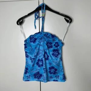 Blå blommig halterneck linnetopp - Supersöt blå linnetopp med mörkblå blommigt mönster och textdetaljer. Toppen har halterneck med knyt och tunna axelband, perfekt för varma dagar. Materialet känns lätt och stretchigt, och toppen har en snygg, figurnära passform.