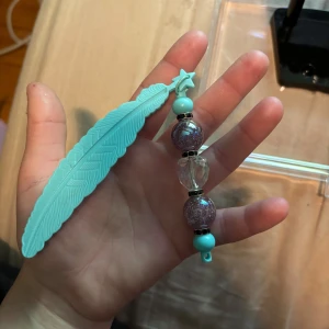 Turkos fjäderformad bokmärke med pärlor - Cool bokmärke med en stor turkos fjäder i plast och glittriga lila pärlor, samt en genomskinlig pärla i mitten. Små turkosa detaljer och en stjärnformad pärla upptill. Perfekt accessoar för att piffa upp väskan eller nyckelknippan.