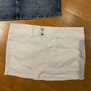 Vit kortkjol med fickor och dubbla knappar - Snygg vit kortkjol med två fickor fram och två bak, samt dubbla silverfärgade knappar i midjan. Kjolen har bälteshällor och klassisk jeanskjolsstil, perfekt för en fräsch och clean look.