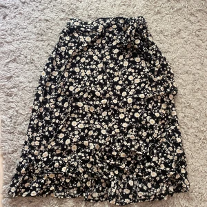 Svart blommig kjol med volang - Supersöt svart kjol med vita och gula blommor och volangkant längst ner. Kjolen har en omlott och knyt i midjan.