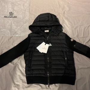 Svart Moncler cardigan  - Svart pufferjacka från Moncler med huva, ribbstickade ärmar och detaljer, samt dragkedja framtill. Jackan har quiltad framsida, två fickor med tryckknappar och Moncler-logga på ärmen. Perfekt för dig som vill ha en clean och stilren look. I nyskick använd några gånger men för liten.  Pris går att diskutera vid snabb affär. Skriv för fler detaljer eller bilder.