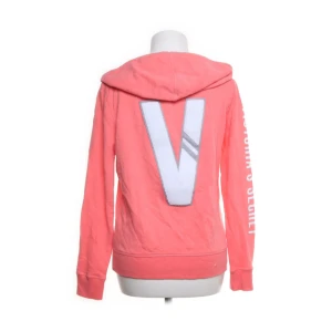 Rosa hoodie från Victoria's Secret - Säljer en snygg rosa hoodie från Victoria's Secret med dragkedja och huva. Stor vit V på ryggen och text på ena ärmen. Mjuk bomullskänsla, perfekt för chill dagar. Fickor framtill och snörning i huvan. Passar dig som gillar streetstyle.