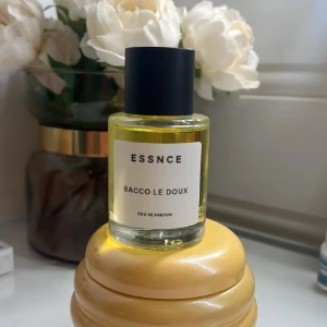 ESSNCE Bacco Le Doux Eau de Parfum - Parfymflaska med minimalistisk design från ESSNCE, doften Bacco Le Doux. Flaskan är cylindrisk i genomskinligt glas med svart matt kork och vit etikett. Innehållet är ljusgult. 