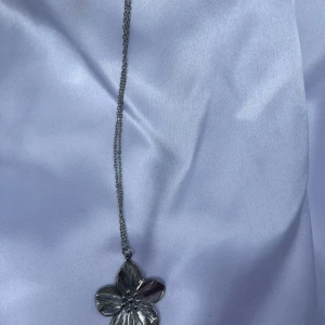Silverfärgat halsband med blomma - Snyggt halsband i silverfärg med en stor, detaljerad blomma som hänge. Kedjan är tunn och smidig, perfekt för att ge din outfit en cool touch. Blomman har fina ingraverade detaljer som gör smycket unikt och stilrent.