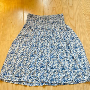 Blommig blå kjol från Indiska - Supersöt blå kjol från Indiska med vitt blommönster och smockad midja. 🩵🤍💙Kjolen har ett otroligt mjukt och skönt material 🥰