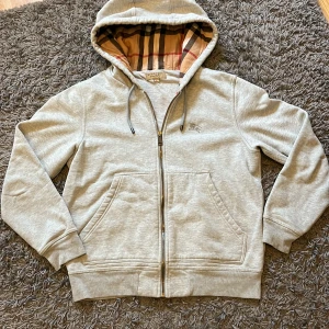 Burberry hoodie - Snygg grå hoodie från Burberry med klassiskt rutigt foder i huvan och broderad logga på bröstet. Tröjan har dragkedja, snörning i huvan och två stora fickor framtill. Perfekt för dig som gillar stilren design med lyxig touch. Bara att skriva vid frågor eller intresse. Pris kan diskuteras😇