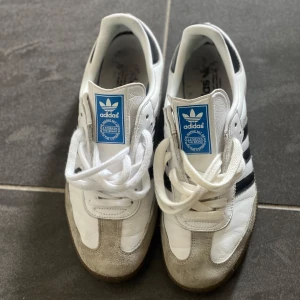 Adidas Samba vita och svarta sneakers - Klassiska Adidas Samba sneakers i vitt skinn med svarta detaljer och grå mocka framtill. Skorna har de ikoniska tre svarta ränderna på sidan, blå logga på plösen och gummisula. Perfekta för dig som gillar retrostil och streetwear.pris kan diskuteras 