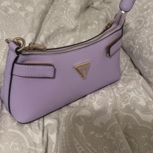 Lila axelväska från Guess med kedja - Superfin lila axelväska från Guess med guldfärgad kedja och dragkedja upptill. Väskan har ett klassiskt Guess-triangelmärke framtill och flera fack inuti för kort och småsaker. Tillverkad i syntetmaterial med stilren design och justerbar axelrem. Aldrig använd 