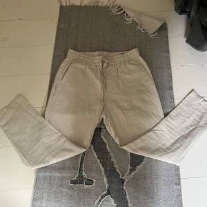Beiga linnebyxor Only & Sons M - Snygga beiga linnebyxor från Only & Sons i modellen Linus. Byxorna har tapered och croppad passform, elastisk midja med snörning och sidfickor. Perfekta för en chill och stilren look under varmare dagar.