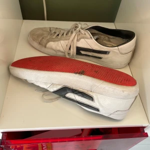 Vita Diesel sneakers med svart detalj - Säljer ett par vita Diesel sneakers i skinn med svarta detaljer på sidorna och röd sula undertill. Skorna har snörning och en klassisk rund tå. Perfekta för dig som gillar streetstyle och designer. Dom är rätt så slitna och lädret är lite skrynkligt men det syns mindre när man har dom på sig. Och det är ett hål därbak 