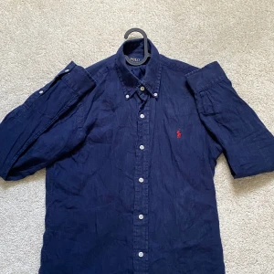 Mörkblå linneskjorta Polo Ralph Lauren - Snygg mörkblå skjorta från Polo Ralph Lauren i slim fit-modell. Tillverkad i linne med klassisk button down-krage, vita knappar och ikonisk röd logga broderad på bröstet. 