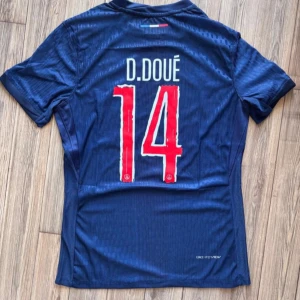 PSG matchtröja D. Doué #14 Nike - Snygg PSG fotbollströja med D. Doué och nummer 14 på ryggen. Tröjan är marinblå med en röd vertikal rand och vita detaljer, officiella klubbmärket och Champions League-märke på ärmen. Tillverkad i lätt och ventilerande material, perfekt för fotbollsälskare.