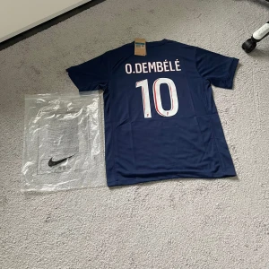 PSG Dembélé 10 Nike fotbollströja - Snygg PSG fotbollströja med O. Dembélé och nummer 10 på ryggen. Tröjan är marinblå med röd och vit rand längs framsidan, PSG-logga på bröstet och Qatar Airways som sponsor. Tillverkad i lätt och ventilerande material, perfekt för match eller träning.