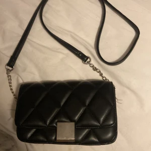 Svart axelväska med kedja - Snygg svart axelväska med silverfärgad kedja Väskan har ett innerfack med dragkedja och lång axelrem i både skinnimitation och kedja. Perfekt storlek för det viktigaste