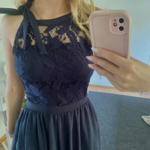 Mörkblå spetsklänning med knyt - Supersnygg mörkblå klänning med spets upptill och knyt vid axeln. Klänningen har en markerad midja och ett flowy kjolparti i tunt tyg. Perfekt för dig som vill sticka ut med en elegant och feminin look. Ärmlös modell med hög halsringning.