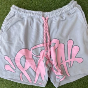 Grå shorts med rosa graffiti-tryck - Syns world set Snygga grå shorts och tröja med stort rosa graffiti-inspirerat tryck framtill och breda, tvinnade snören i midjan. Mjukt bomullsmaterial och avslappnad passform, perfekta för chill dagar eller streetwear-stil.