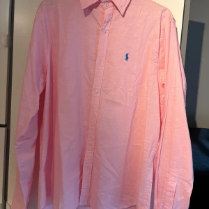 Rosa skjorta från Ralph Lauren L - Snygg rosa skjorta från Ralph Lauren i storlek L. Klassisk modell med knappar framtill, button down-krage och blå broderad logga på bröstet. Tillverkad i mjuk bomull och har långa ärmar. Perfekt för dig som vill ha en stilren och fräsch look.