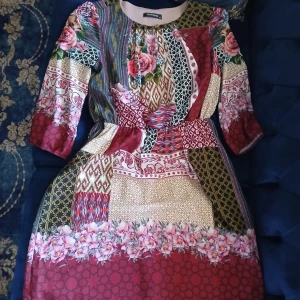 Färgglad blommig klänning från Myrvang - Unik klänning från Myrvang i storlek 40 med färgstarkt patchworkmönster, blommiga detaljer och inslag av vinrött, beige, grönt och rosa. Modellen har trekvartsärm med resår och rund halsringning. Perfekt för dig som gillar statement-plagg!