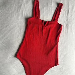 Röd ribbad baddräkt med v-ringning - Snygg röd body med breda axelband och en subtil v-ringning framtill. Materialet är ribbat vilket ger en cool struktur och extra stilpoäng. 